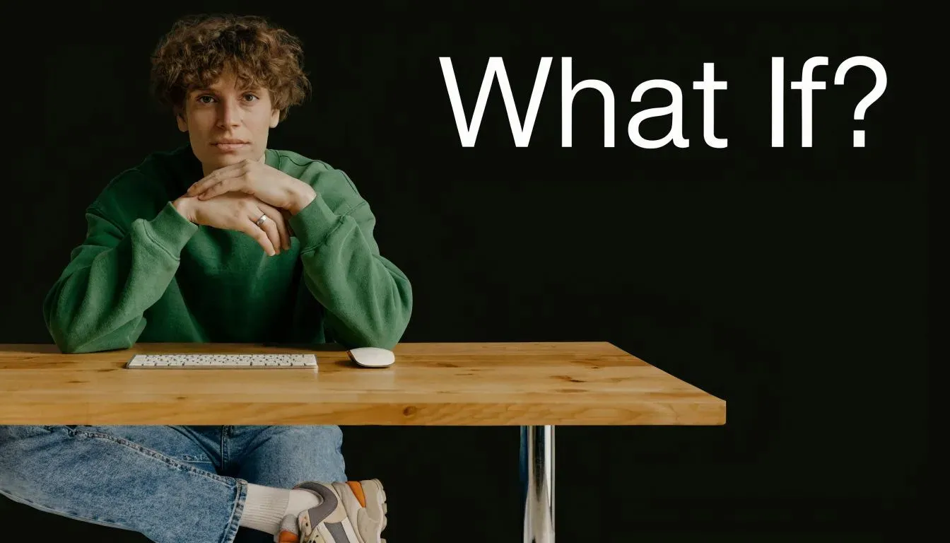 A young man with curly hair wearing a green sweater sits at a desk with a computer keyboard.